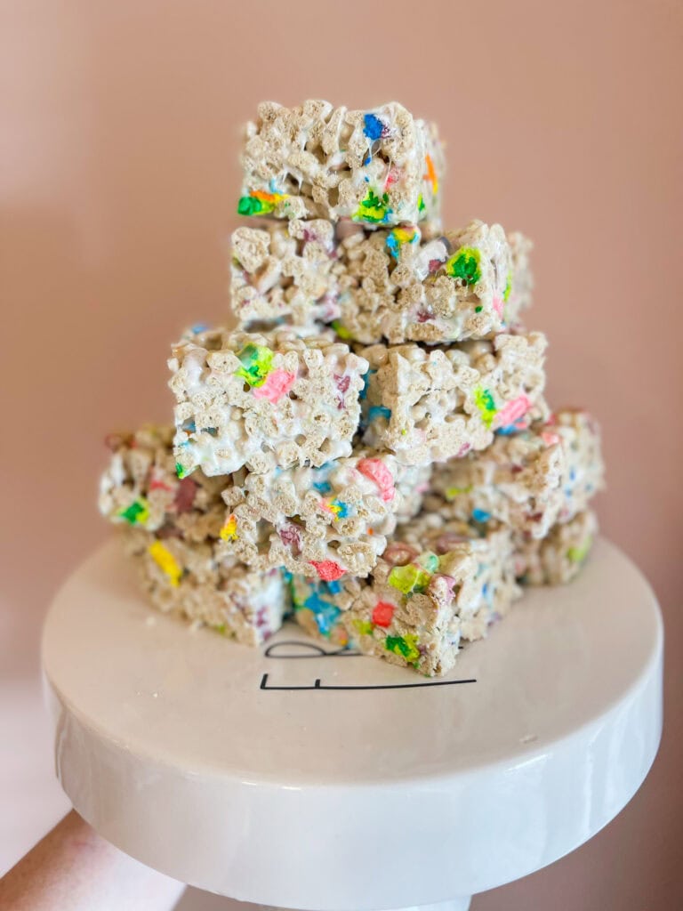 A stack of colorful cereal treats, made with marshmallows and rainbow cereal pieces, sits on a white cake stand against a pale pink background.