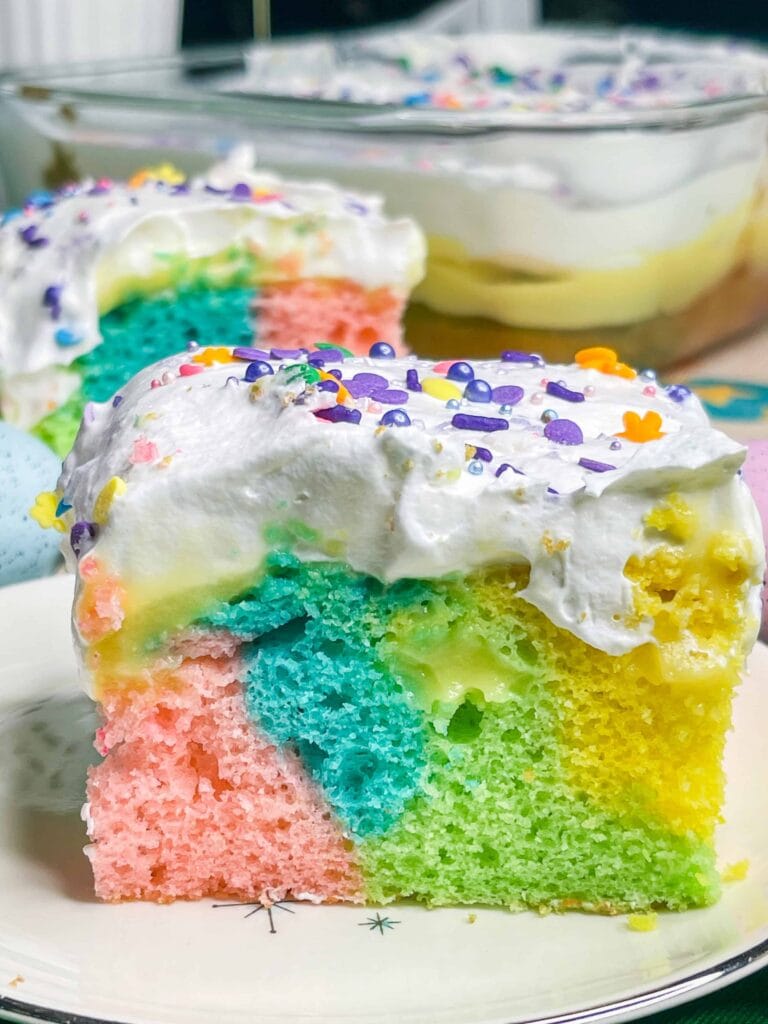 A slice of colorful, pastel rainbow cake with layers of pink, blue, green, and yellowโperfect for a St. Patrickโs Day Treat Ideas Roundupโis topped with whipped cream and sprinkles and served on a white plate. Another slice sits in the background.