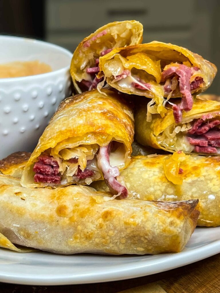 A plate of baked egg rolls filled with sliced corned beef, sauerkraut, and melted cheese, stacked next to a white bowl of dipping sauce. The egg rolls are golden brown and crispy.