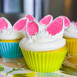 Three cupcakes with white frosting and bunny ear decorations made of pink and white marshmallows sit on a floral plateโperfect for a St. Patrickโs Day Treat Ideas Roundup. The cupcakes are in colorful silicone wrappers: blue, green, and yellow.