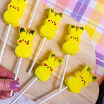 A hand holds a lollipop stick with a yellow marshmallow shaped like a Pikachu face. Several similar Pikachu marshmallow pops are arranged on a wooden boardโperfect for any St. Patrickโs Day Treat Ideas Roundupโover a pastel plaid tablecloth.