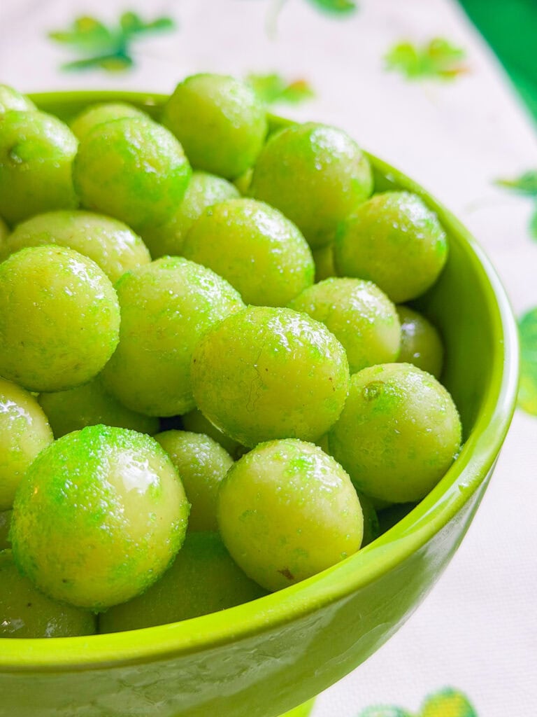 A green bowl filled with small, round, shiny green candies or snacks, with a few more visible in the background on a white surface decorated with green patterns.