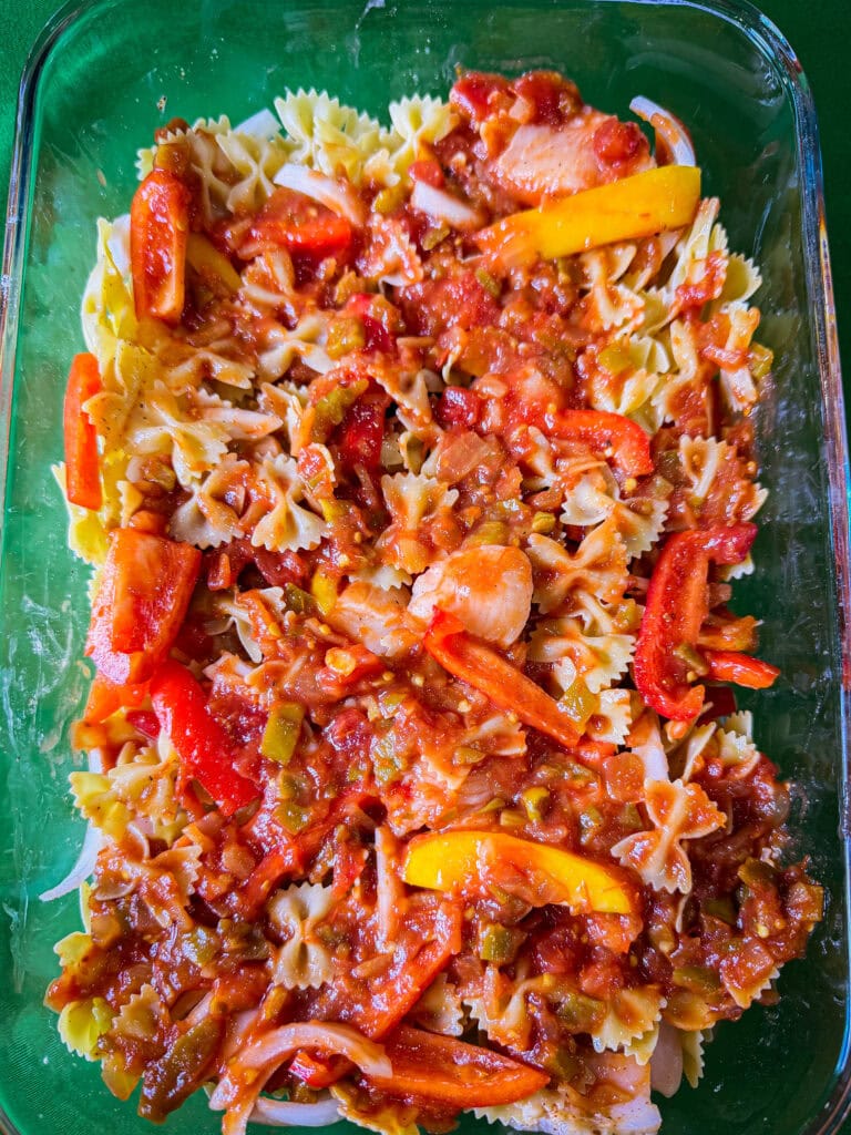 A glass baking dish filled with bow-tie pasta topped with tomato sauce, sliced red and yellow bell peppers, onions, and mixed vegetables. The dish is colorful and appears ready to be baked or served.
