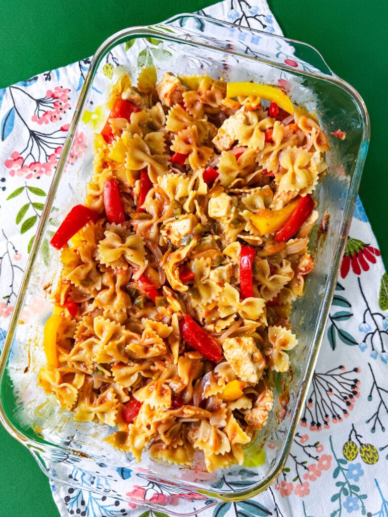 A glass baking dish filled with bowtie pasta, chunks of chicken, and sliced red and yellow bell peppers sits on a colorful floral-patterned cloth over a green surface.