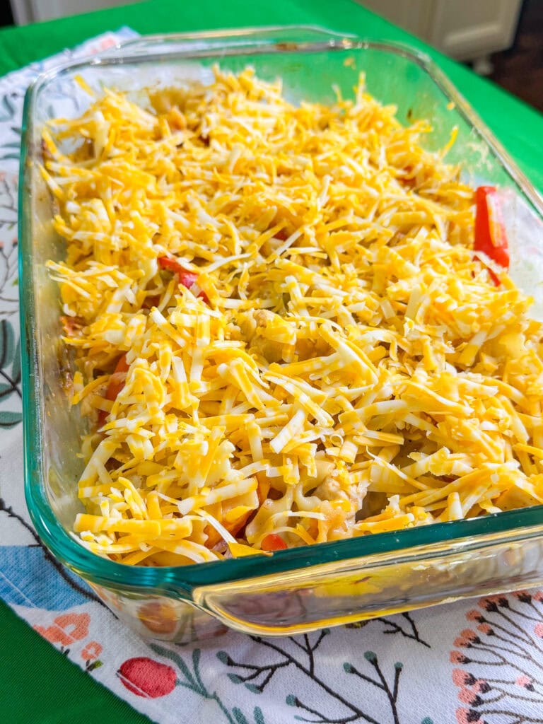 A glass baking dish filled with a casserole topped with a generous layer of shredded yellow and white cheese, resting on a colorful patterned cloth on a green surface.
