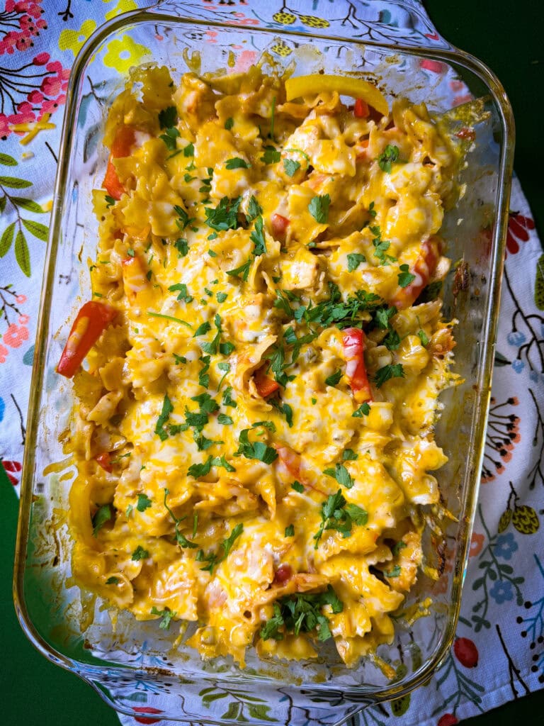 A glass baking dish filled with cheesy baked pasta, topped with melted cheese and chopped herbs, sits on a patterned cloth with colorful floral designs.