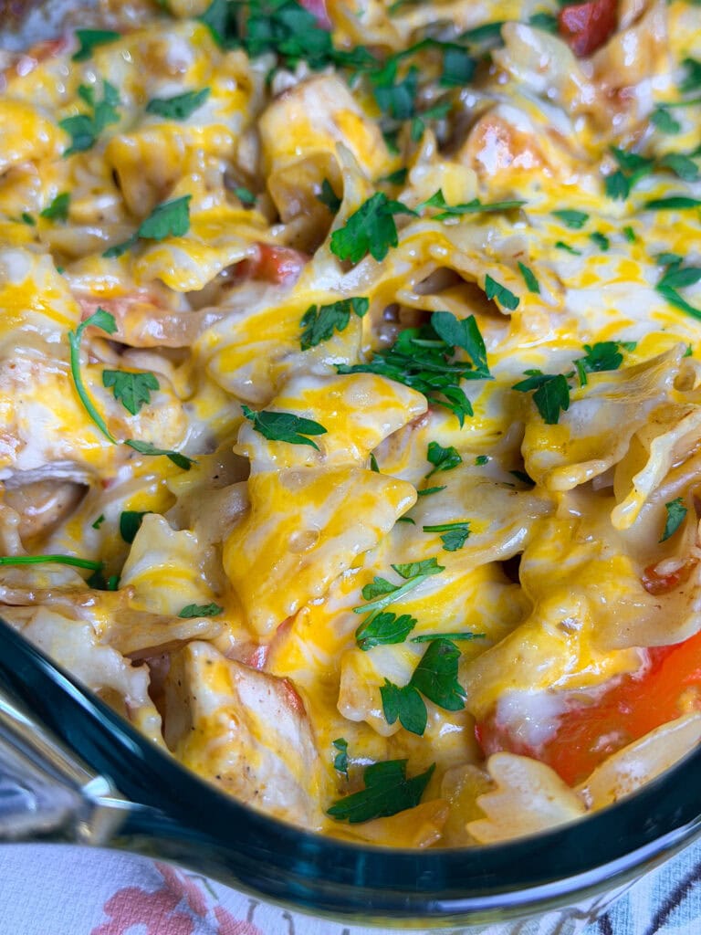 Close-up of a cheesy casserole topped with melted cheese, chopped parsley, and visible chunks of chicken, pasta, and red bell peppers in a glass baking dish.