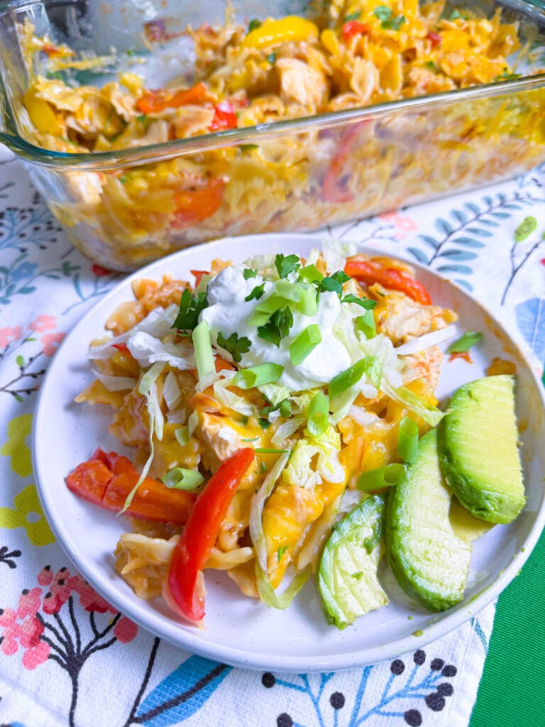A plate of cheesy chicken casserole with sliced red peppers, shredded lettuce, scallions, sour cream, and avocado slices, served in front of a casserole dish on a floral tablecloth.