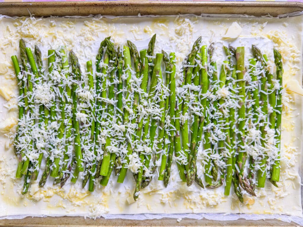 A rectangular pastry topped with a layer of cream, fresh asparagus spears arranged in rows, and sprinkled with shredded cheese and pieces of butter, ready to be baked.