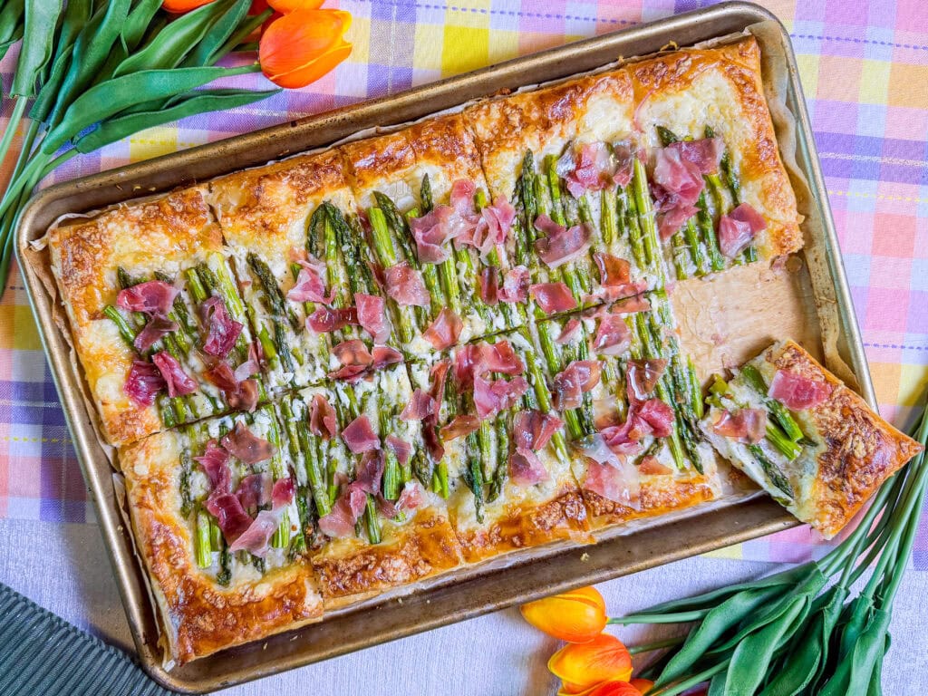 A rectangular puff pastry tart topped with asparagus, prosciutto, and cheese, sliced into squares on a baking sheet, surrounded by orange tulips on a colorful plaid tablecloth.