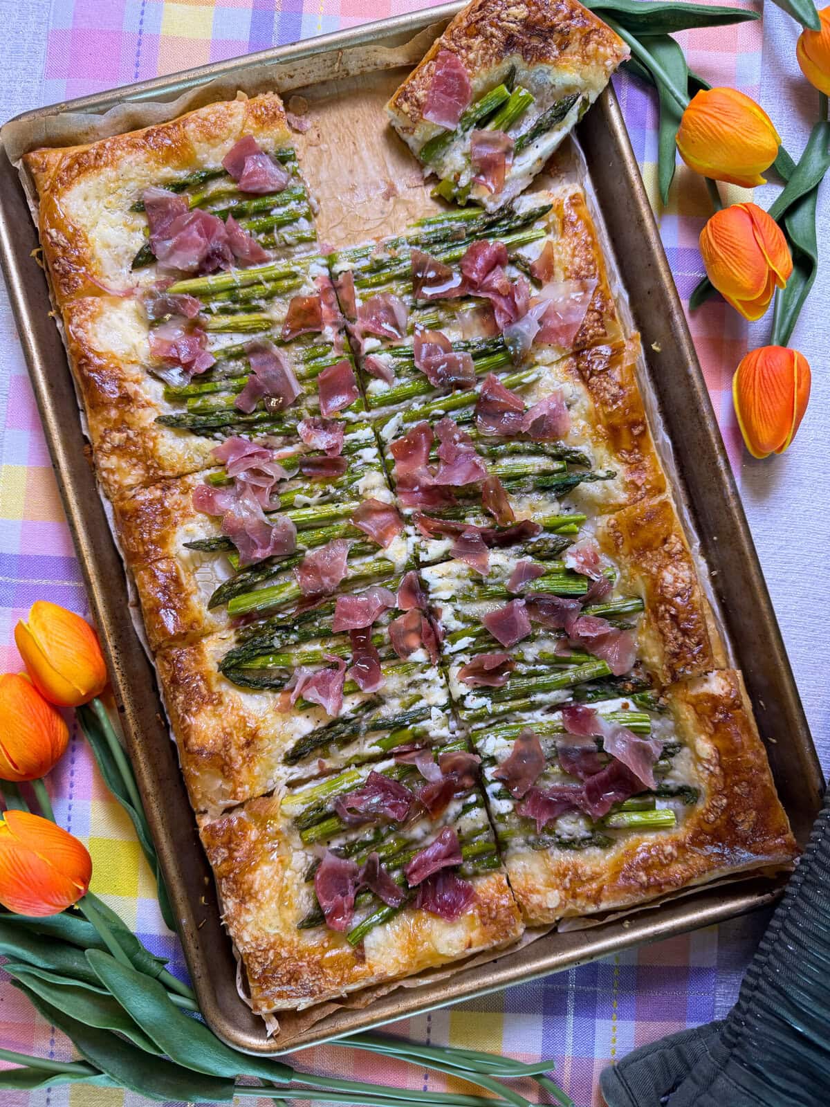 A rectangular puff pastry tart topped with asparagus, melted cheese, and pieces of prosciutto, sliced into squares on a baking sheet. Orange tulips and a pastel plaid tablecloth decorate the scene.