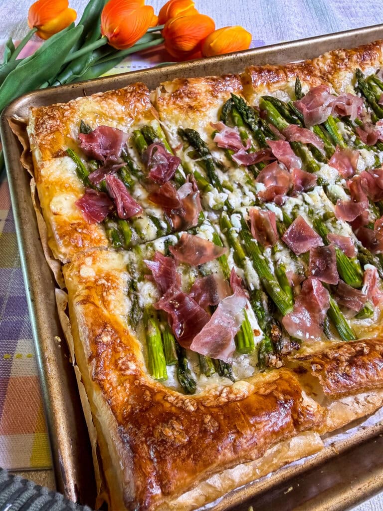 A golden puff pastry tart topped with asparagus, crumbled cheese, and pieces of prosciutto sits on a baking tray. Orange tulips and a colorful plaid cloth are in the background.