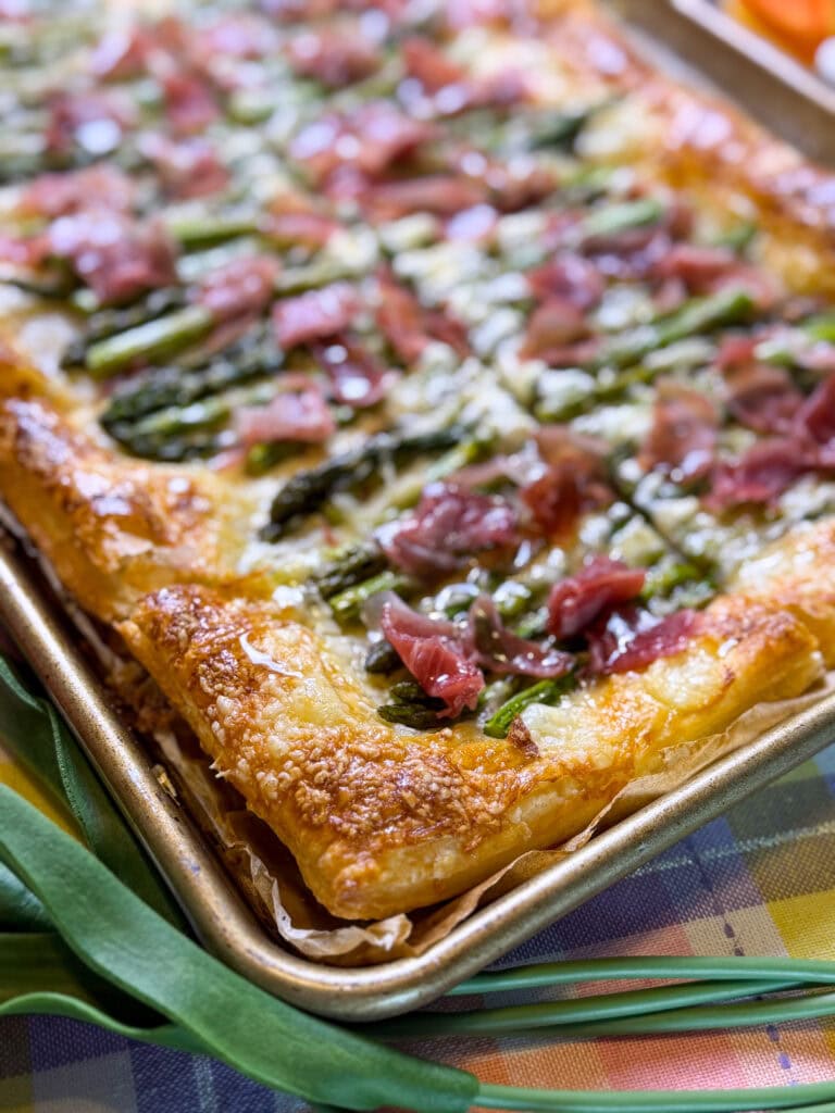 A close-up of a golden, flaky puff pastry tart topped with asparagus, prosciutto, and cheese, served on a baking sheet. The tart is set on a colorful, checkered tablecloth.