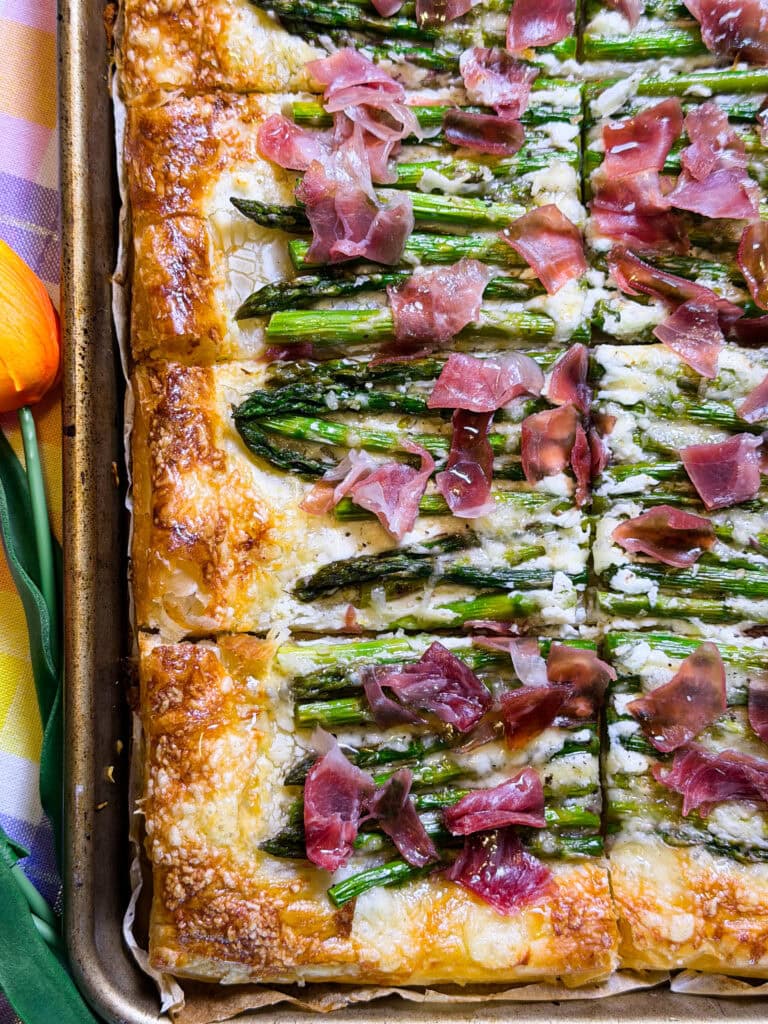 A close-up of a baked puff pastry tart topped with asparagus, melted cheese, and pieces of prosciutto, sliced into squares on a baking sheet.