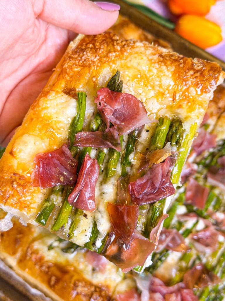 A close-up of a hand holding a slice of focaccia topped with asparagus, melted cheese, and thin slices of prosciutto. The background shows more of the focaccia and some colorful vegetables.