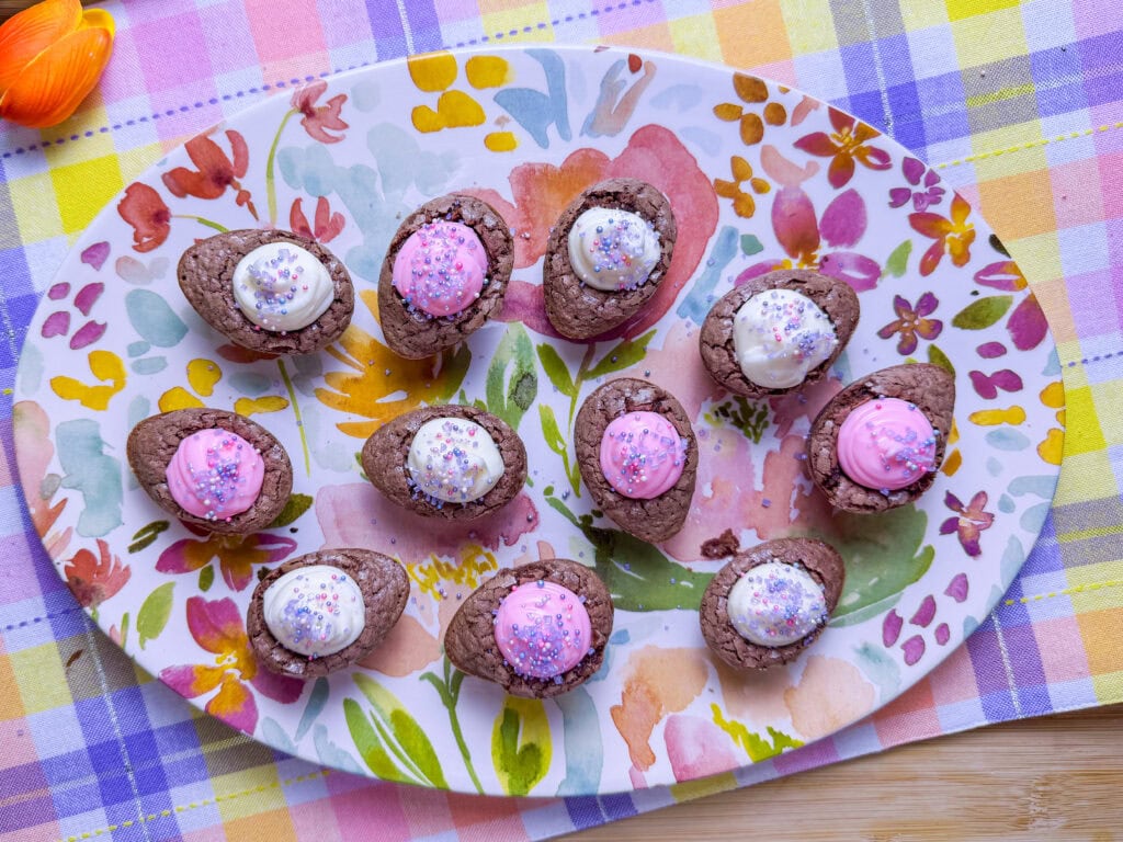 A floral platter holds chocolate egg-shaped treats, each topped with pink or white frosting and colorful sprinklesโa delightful addition to any St. Patrickโs Day Treat Ideas Roundupโset on a pastel plaid tablecloth.