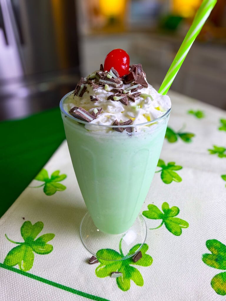 A green milkshake in a clear glass topped with whipped cream, chocolate shavings, and a cherry, with a green straw, sits on a white tablecloth decorated with green shamrocks.