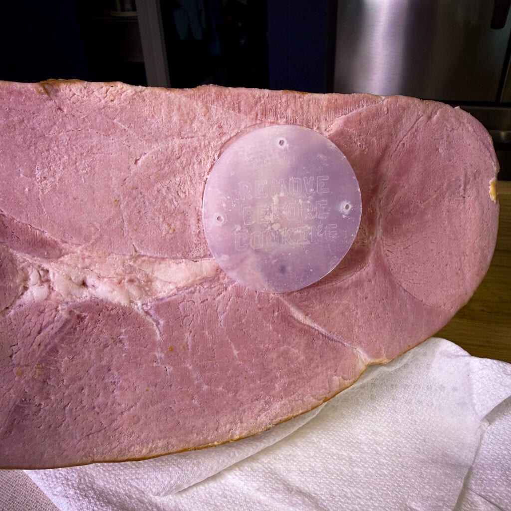 A close-up of a sliced ham shows a round, translucent plastic disc embedded in the surface with raised text that reads REMOVE PLASTIC BONE COVER. The ham rests on a white paper towel.