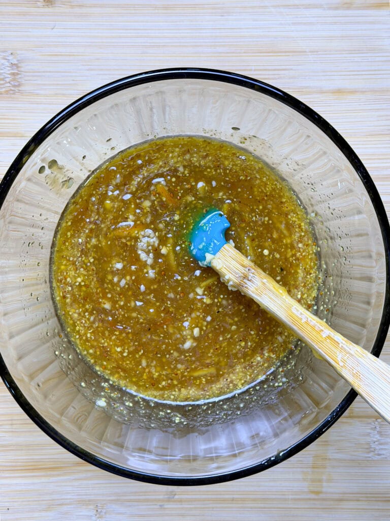 A glass bowl filled with a golden, slightly chunky liquid mixture and a blue silicone spatula with a wooden handle, resting on a light wooden surface.