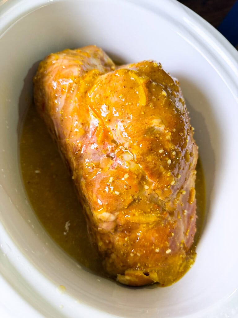 A seasoned, marinated pork roast sits in a white slow cooker, ready to be cooked. The meat is coated with a yellowish sauce and visible spices.