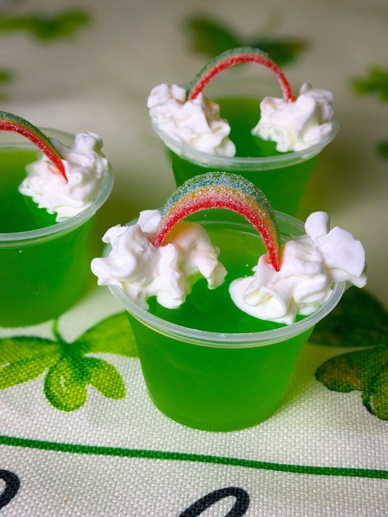 Three small clear cups filled with bright green gelatin are topped with whipped cream and a rainbow-colored candy strip shaped like an arch. The cups are placed on a surface decorated with green shamrocks.