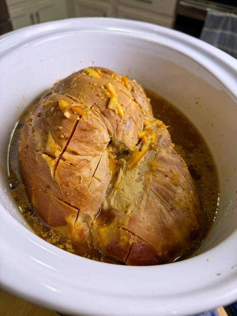A cooked ham with scored lines sits in a white slow cooker, surrounded by broth and pieces of melted seasoning or cheese, in a kitchen setting.
