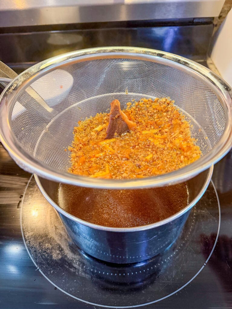 A fine mesh strainer holding a mixture of spices and orange peel rests over a glass bowl, filtering a dark liquid on a kitchen stovetop.
