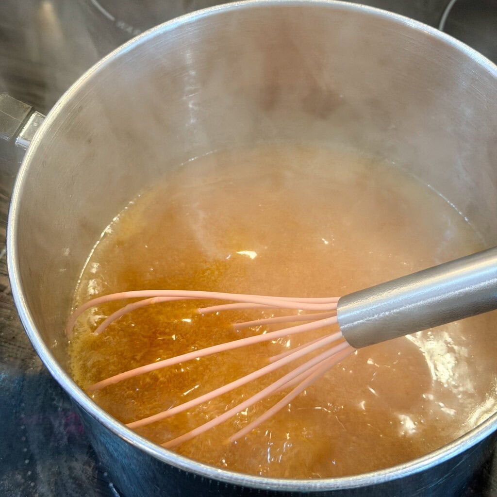 A metal pot filled with a bubbling, amber-colored liquid is being stirred with a pink whisk. Steam rises from the hot mixture, indicating it is being cooked on a stove.