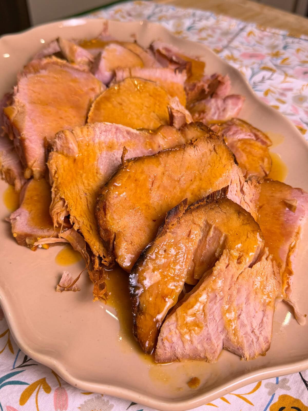 A platter of sliced ham with a golden-brown glaze, arranged on a light-colored serving dish atop a patterned tablecloth.