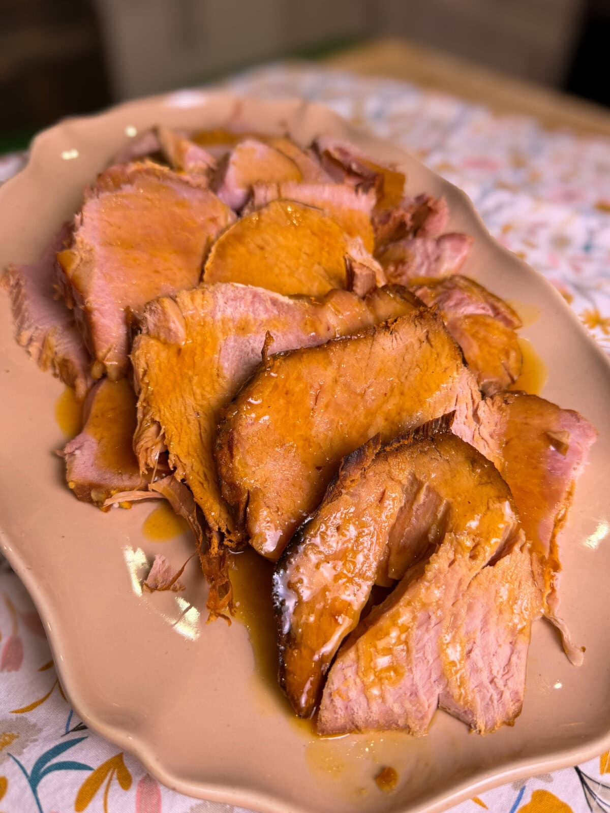 Sliced roast pork with a golden-brown glaze is arranged on an oval beige platter, sitting on a table with a floral-patterned tablecloth.