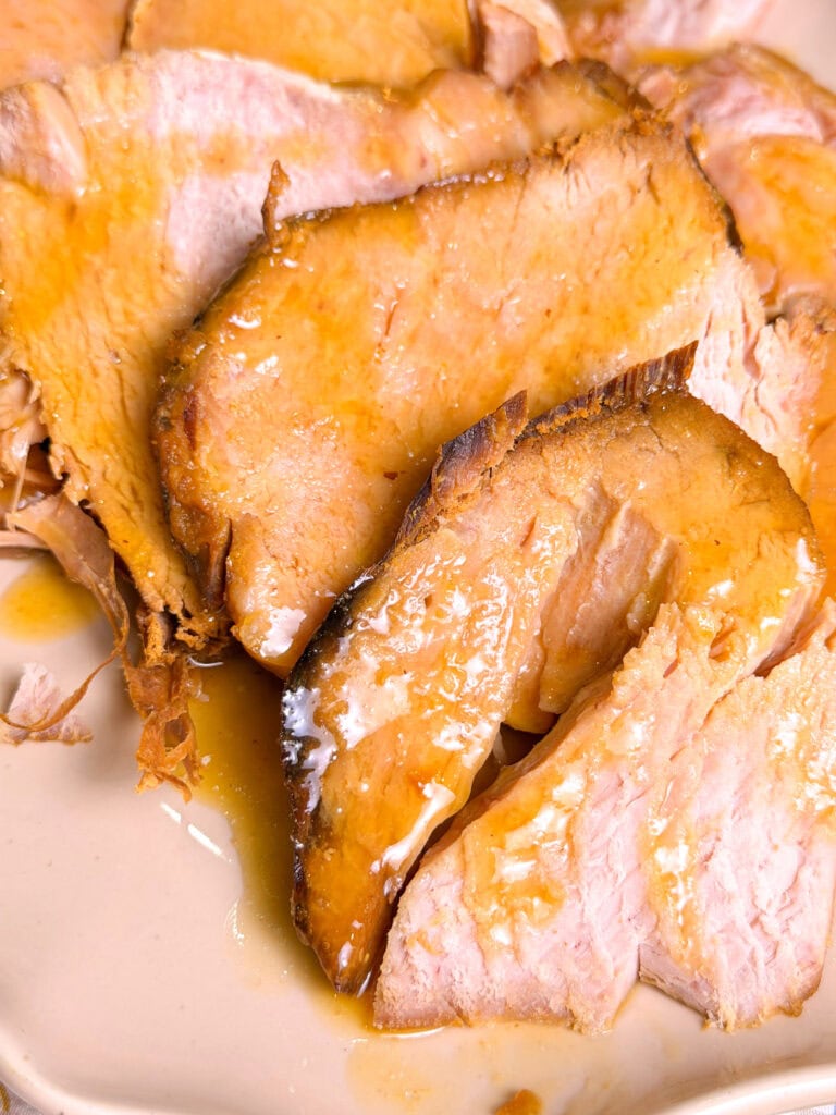 Close-up of several slices of roast pork with golden-brown edges, served on a light-colored plate and topped with a glossy, savory sauce.