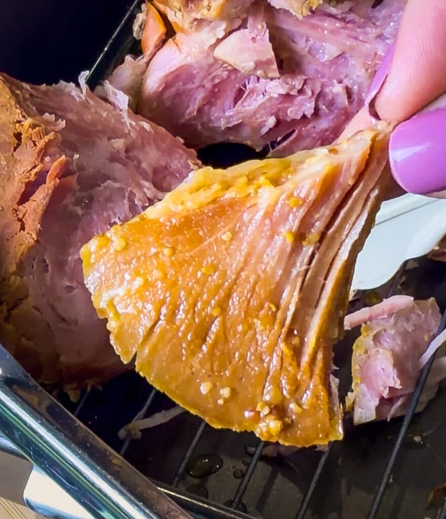 Close-up of a hand with purple nail polish pulling apart a juicy, cooked piece of ham with a golden, seasoned crust. The ham is sitting on a rack inside a tray.