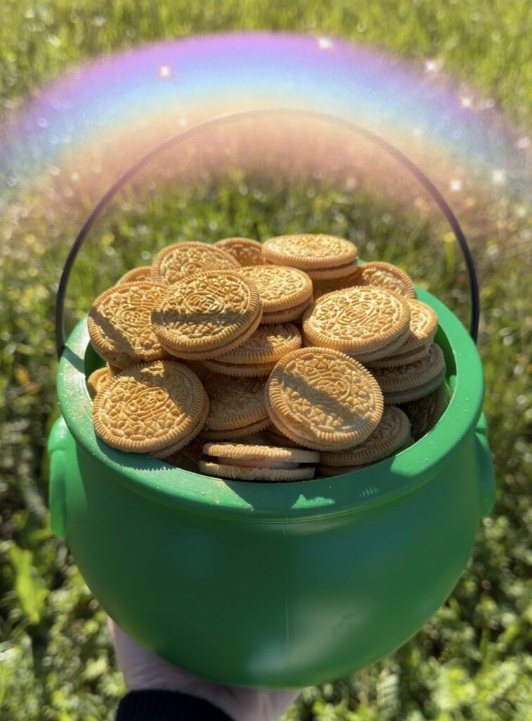 A green plastic pot filled with golden sandwich cookies is held up outdoors, with grass in the background and a bright rainbow arching overhead.