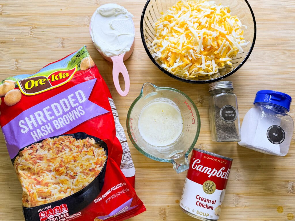 A wooden surface displays ingredients: a bag of shredded hash browns, a measuring cup of sour cream, a bowl of shredded cheese, a measuring cup with melted butter, a can of cream of chicken soup, pepper, and salt containers.