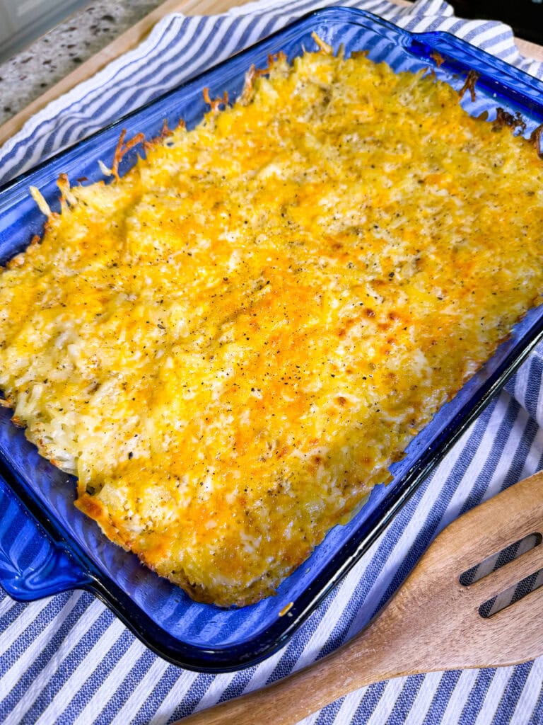 A blue baking dish filled with a golden-brown, cheesy hashbrown casserole sits on a striped kitchen towel next to a wooden slotted spoon.