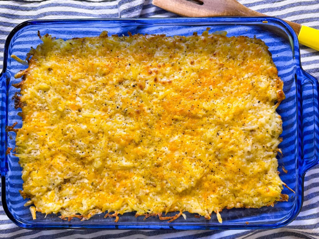 A casserole dish filled with a baked cheesy hash brown casserole, topped with melted golden cheese and black pepper, sits on a blue striped cloth next to a wooden spatula.