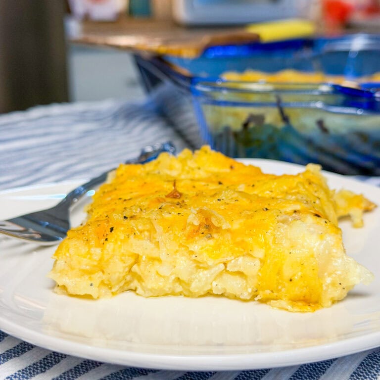 A close-up of a cheesy hash brown casserole served on a white plate with a fork. The casserole is golden brown and topped with melted cheese, with a baking dish of more casserole in the background.