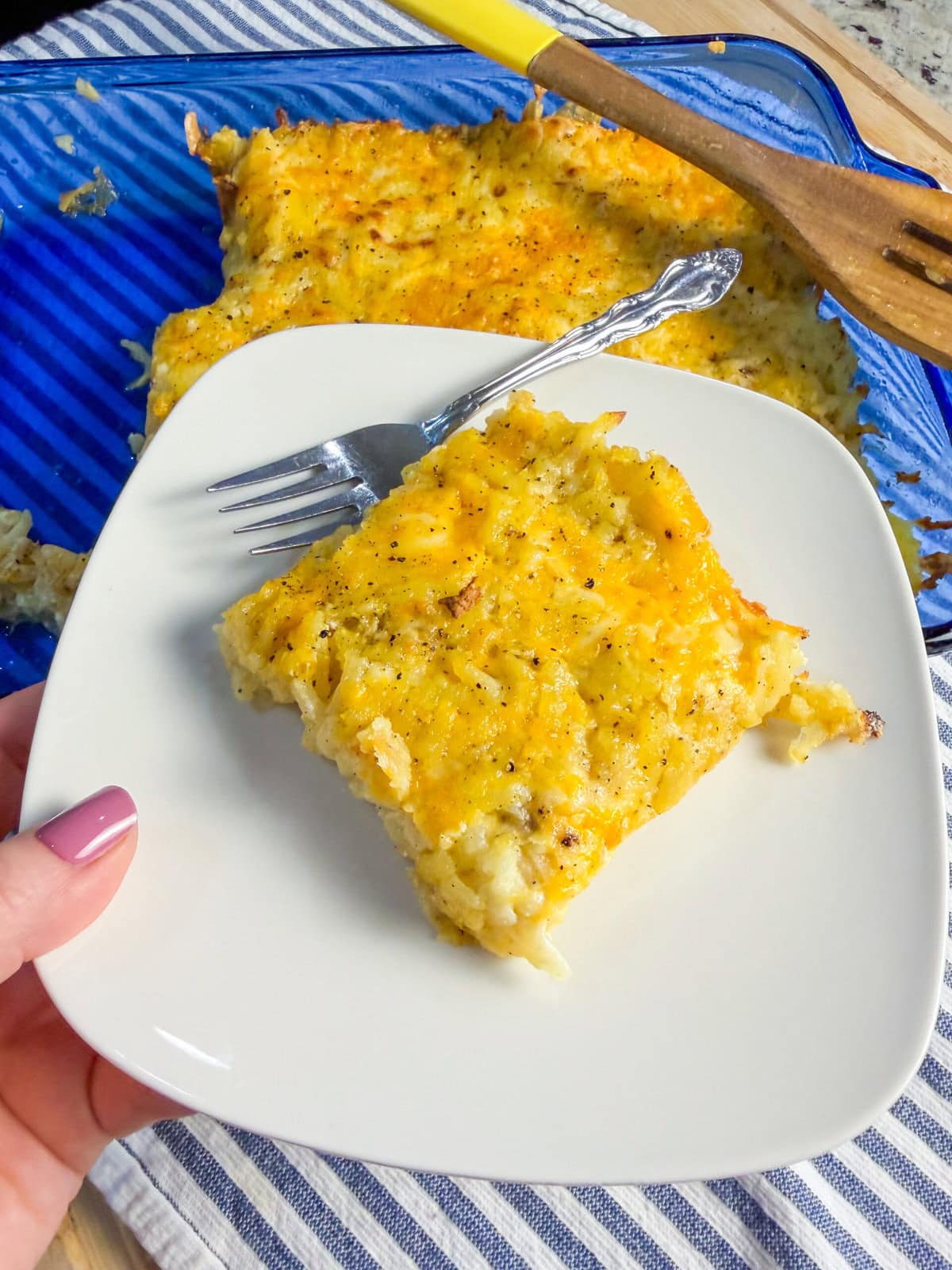 A hand holds a white plate with a square serving of cheesy hash brown casserole and a fork. The casserole dish and a wooden spatula are in the background on a striped cloth.