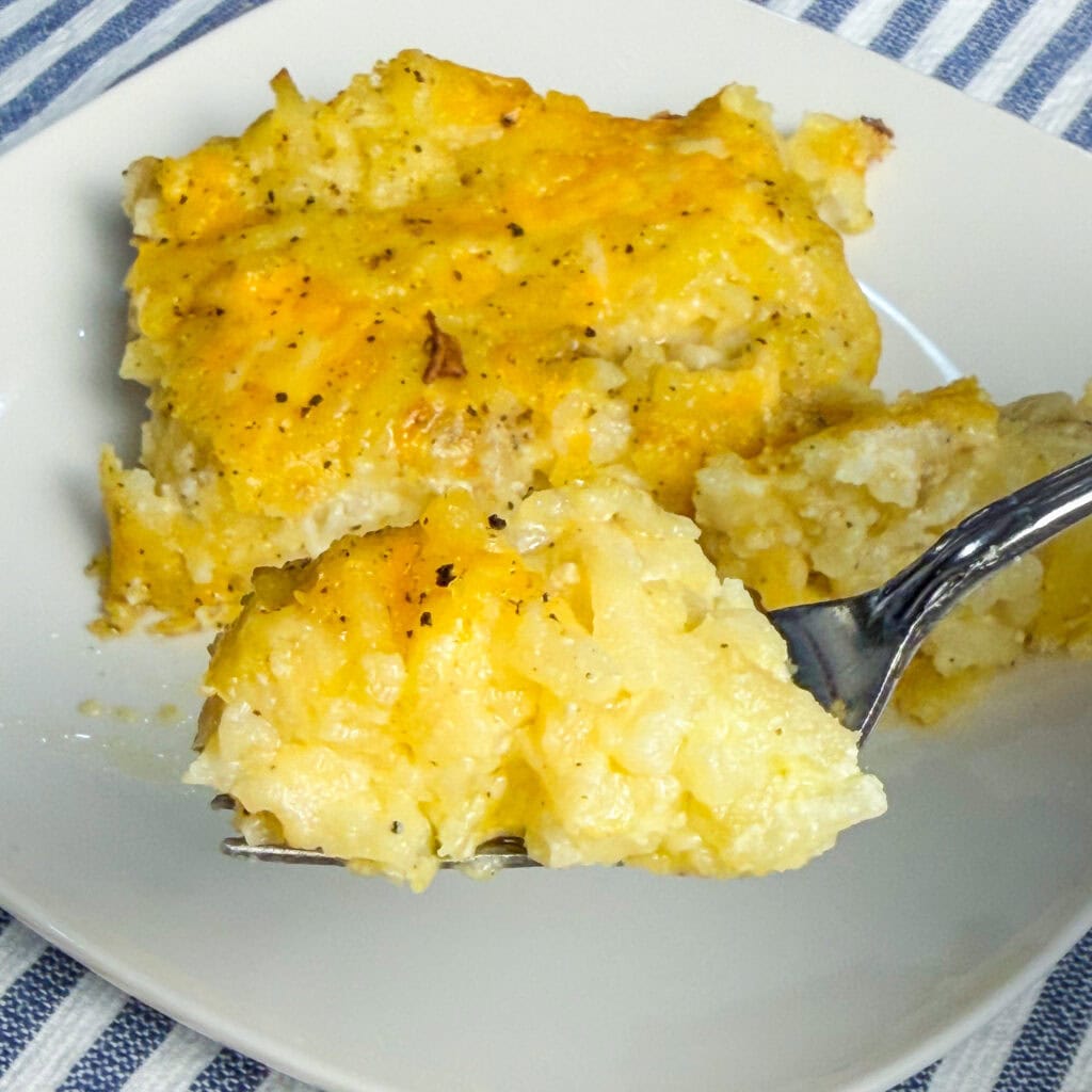 A fork holds a bite of cheesy, baked potato casserole above a plate with a larger serving, topped with melted cheese and black pepper. A blue and white striped cloth is underneath the plate.