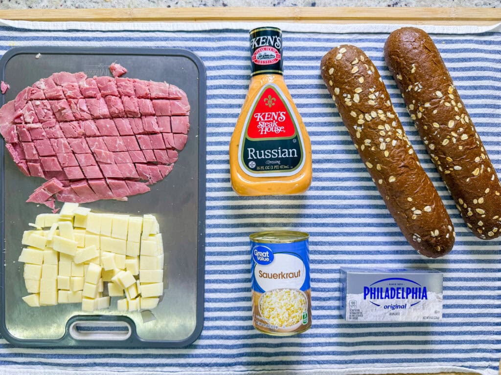 Top-down view of sliced deli meat, cubed cheese, a bottle of Russian dressing, a can of sauerkraut, a package of cream cheese, and two oat-topped sandwich rolls on a blue and white striped towel.