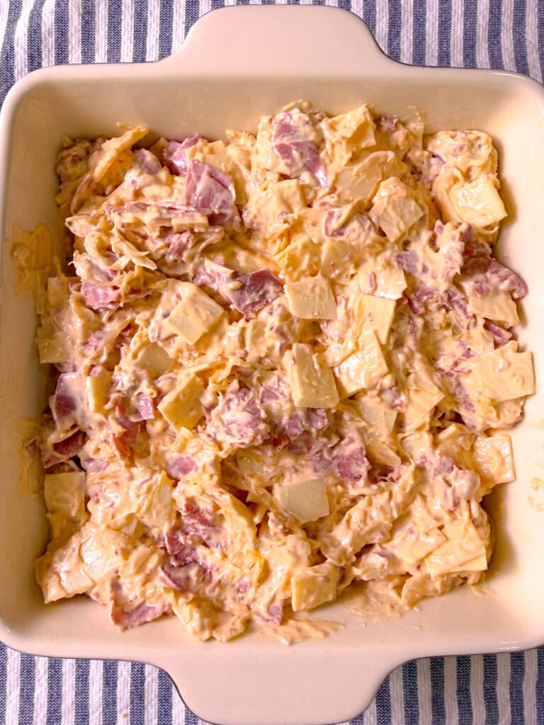 A baking dish filled with a creamy mixture of diced ham, pineapple chunks, and shredded cheese, ready to be baked, sits on a blue and white striped cloth.