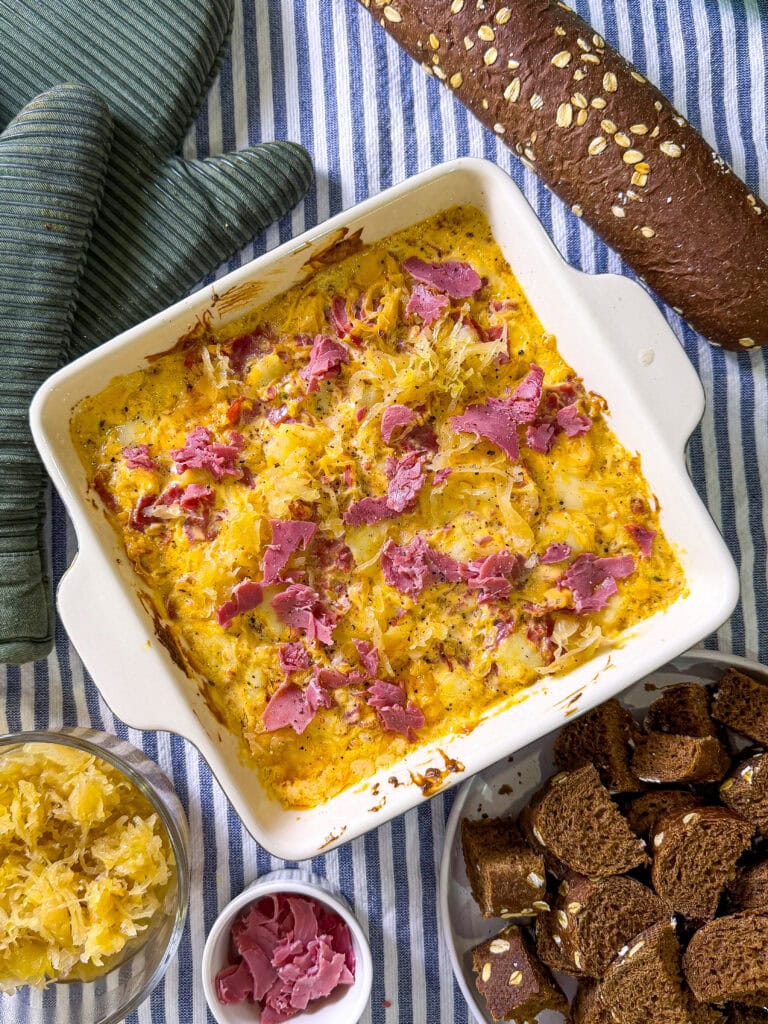 A baked casserole topped with shredded cheese and bits of corned beef sits on a striped cloth, surrounded by dark bread, bread cubes, a jar of sauerkraut, and small bowl of pickled onions.
