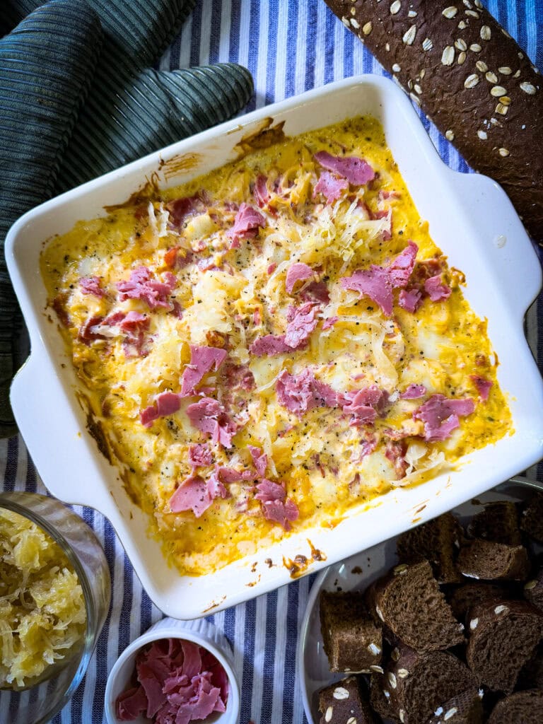 A square baking dish filled with a cheesy baked dip topped with shredded meat sits on a striped cloth, surrounded by a loaf of dark bread, bread cubes, sauerkraut, and a small bowl of pink pickled onions.