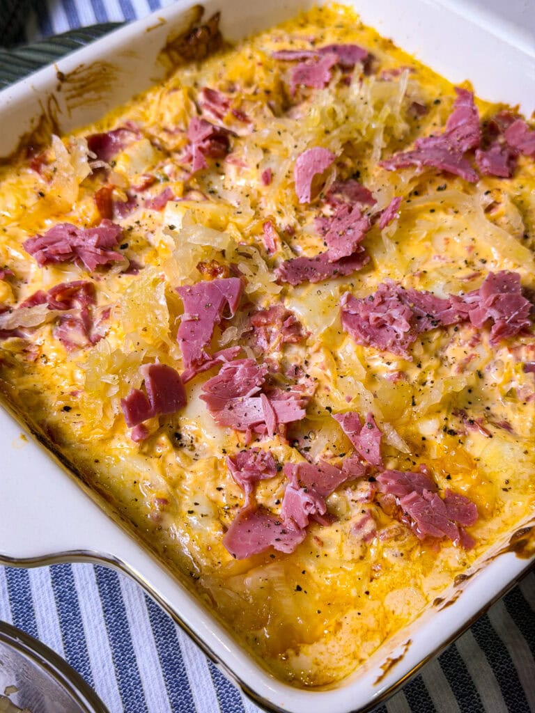 A baked casserole dish filled with a creamy, cheesy mixture topped with shredded corned beef and sauerkraut, resting on a blue-and-white striped cloth.