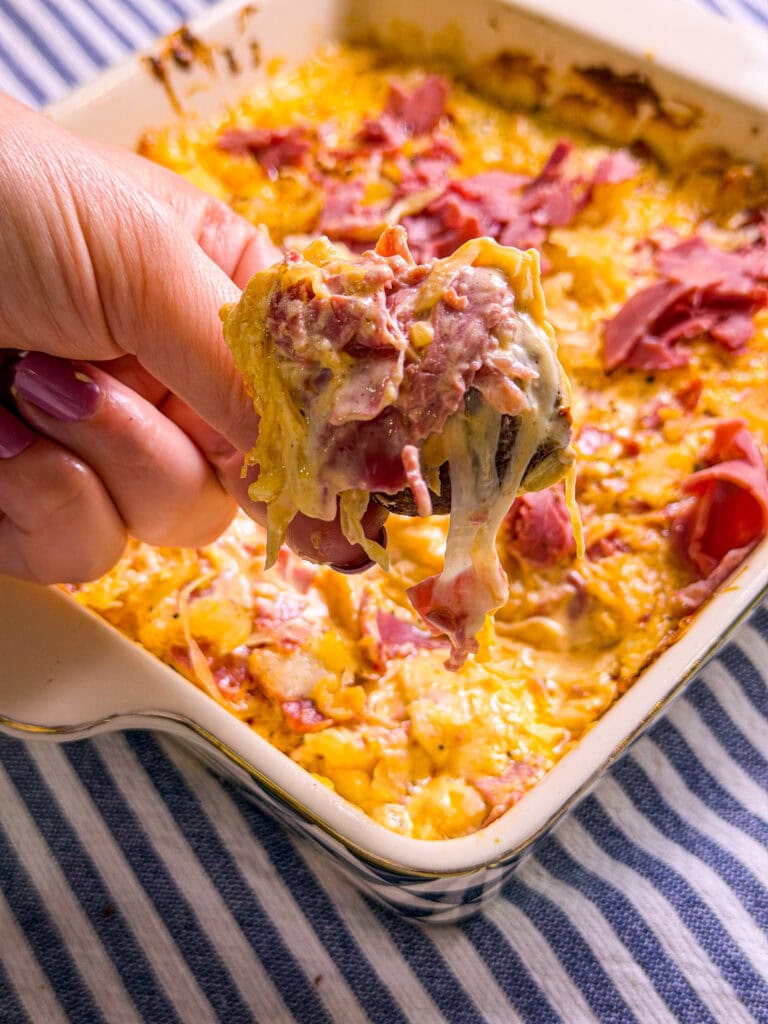 A hand holding a cheesy serving of casserole with shredded meat, above a baking dish filled with the same casserole, on a blue and white striped cloth.