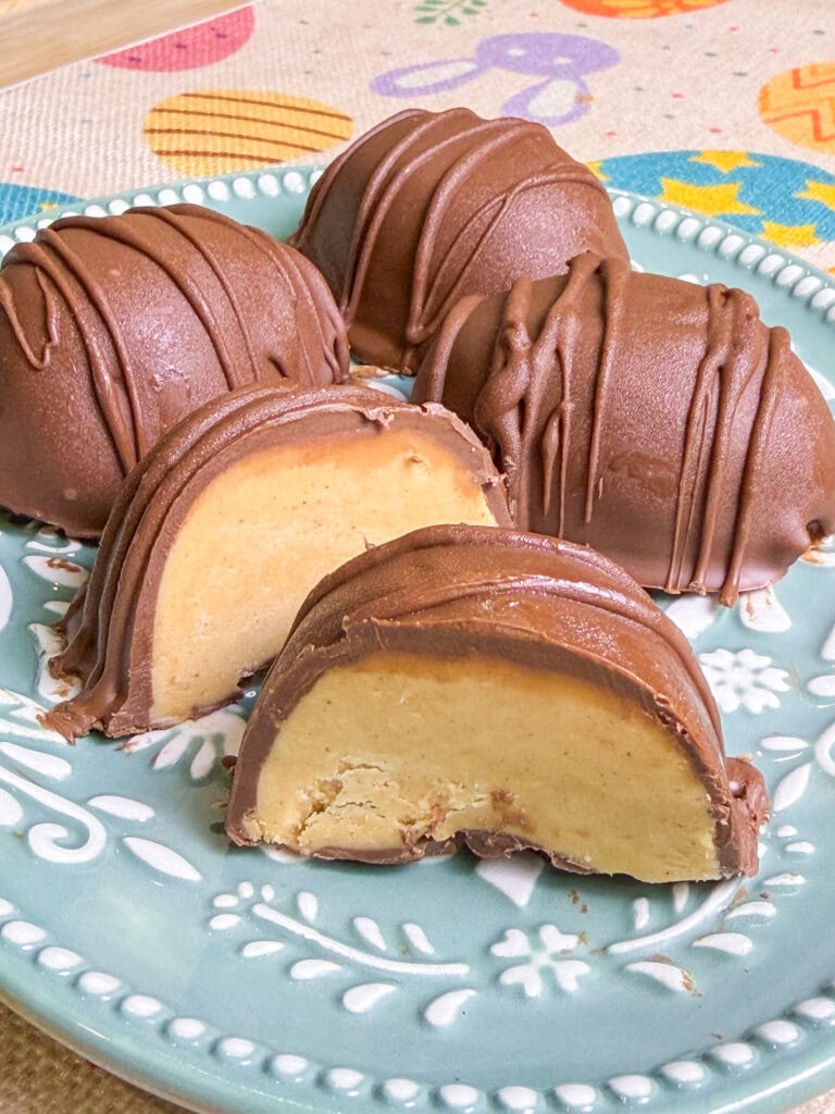 Chocolate-covered peanut butter eggs on a decorative plate, with one cut in half to reveal the creamy filling. The colorful, festive background makes these perfect for any celebration, and a great addition to your St. Patrickโs Day Treat Ideas Roundup.