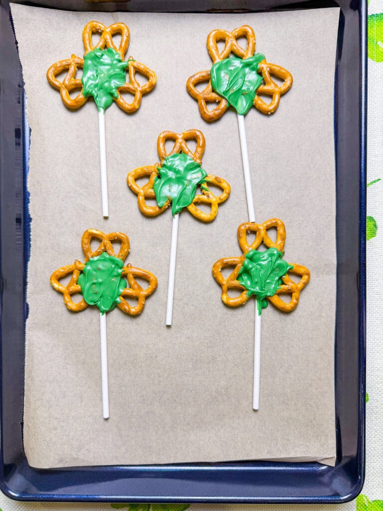 Four shamrock-shaped treats made from pretzels and green icing on sticks are arranged on a parchment-lined baking tray, making them perfect for any St. Patrick’s Day Treat Ideas Roundup.