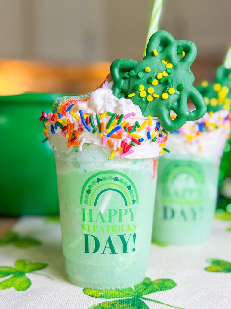 A green frosty drink in a cup labeled Happy St. Patrick's Day, topped with whipped cream, rainbow sprinkles, a green cookie, and a straw sits on a shamrock-patterned tablecloth—perfect for any St. Patrick’s Day Treat Ideas Roundup.