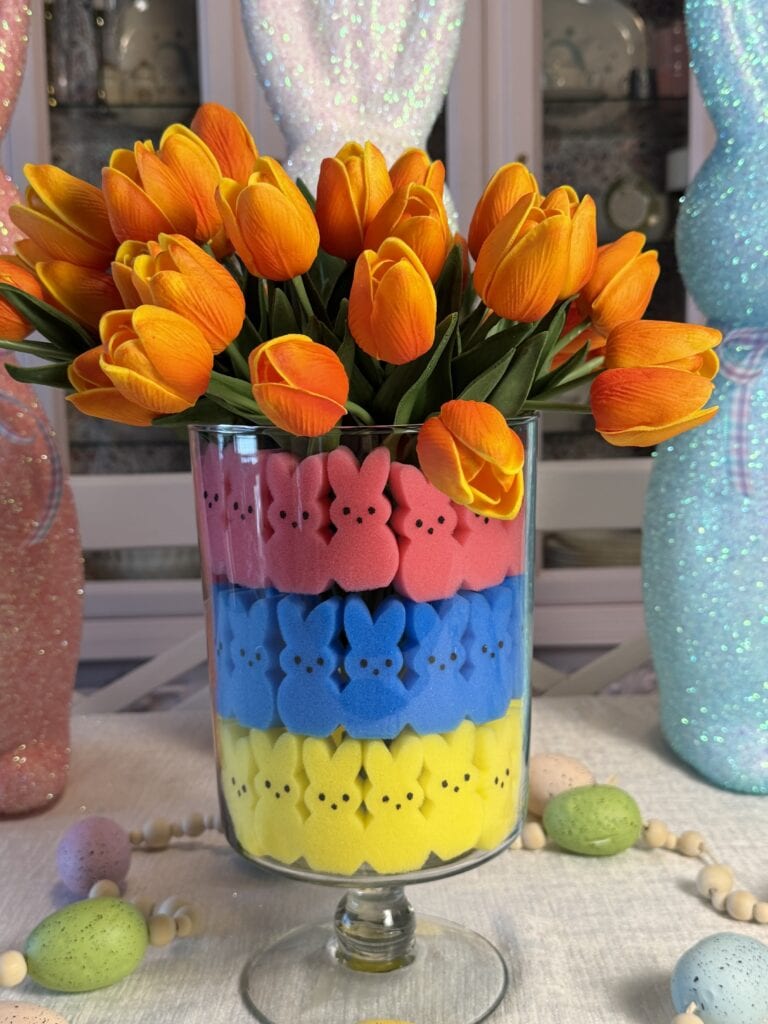 A glass vase holds orange tulips and layers of marshmallow bunny candies in pink, blue, and yellowโan Easy Easter Treat Idea. Decorative eggs and beads scatter the table, with pastel bunny figures in the background.