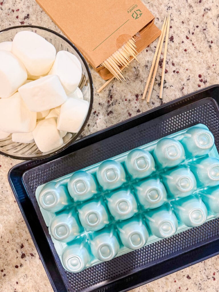 A bowl of large marshmallows, a box of wooden skewers, and a blue egg carton tray are arranged on a kitchen countertop.
